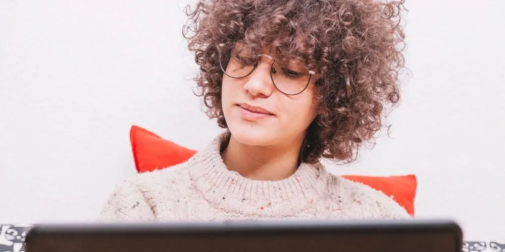 Woman with curly hair and round glasses looking down at a laptop screen, with a red pillow behind her.
